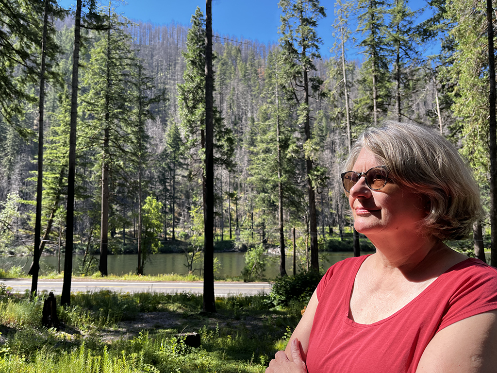 White woman with cropped sandy gray hair stands with arms crossed, in front of a river, nearby the area is green, up on the hillside, trees are dead and grayed.