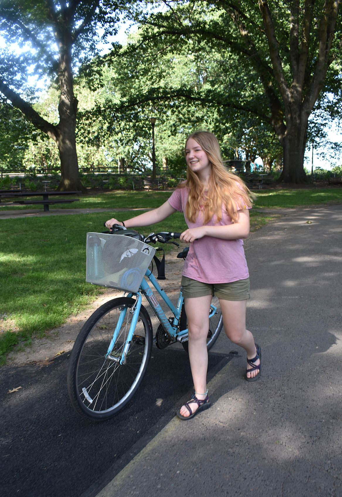 A Teenager walks a bike with a basket on a park walkway.
