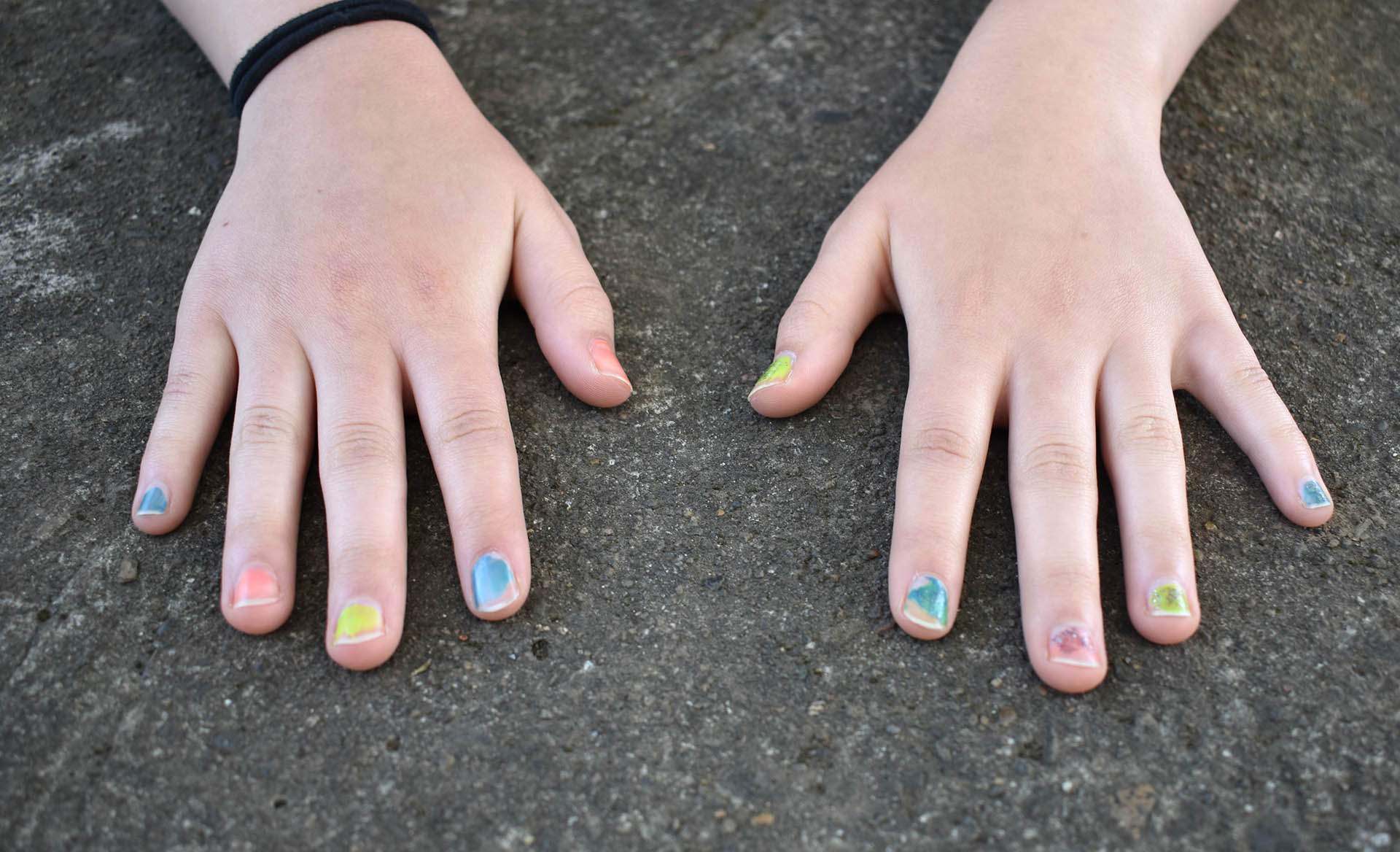 Two hands rest on a concrete table, featuring multi-colored nails and hairbands around the young girl’s wrist.