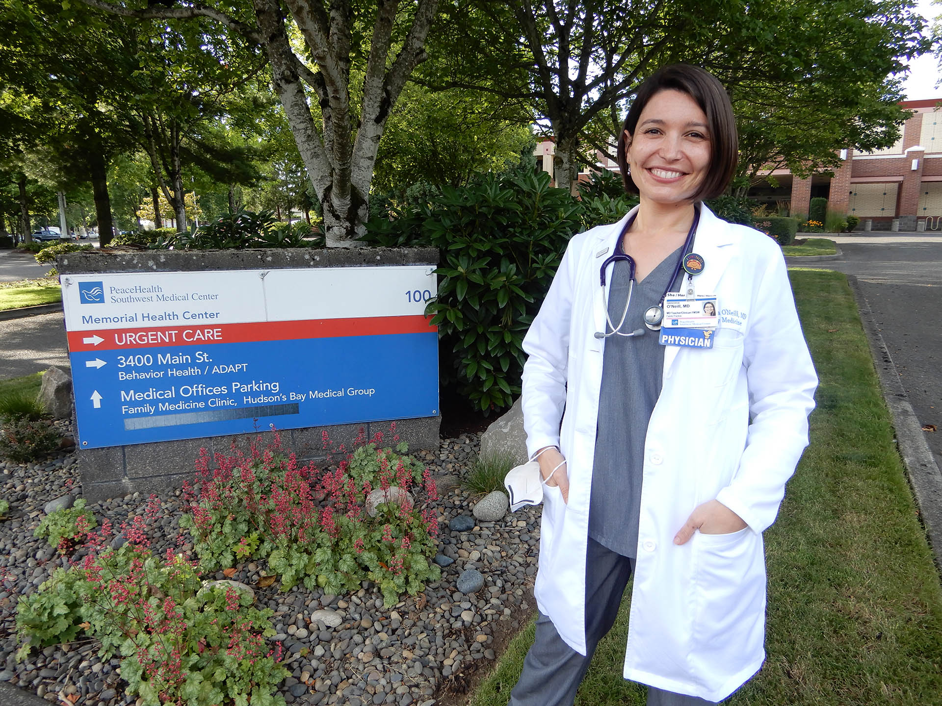 A female Latina physician in a white coat stands in front of a sign for PeaceHealth Family Medicine of Southwest Washington Center, a family health clinic in Vancouver, Wash.