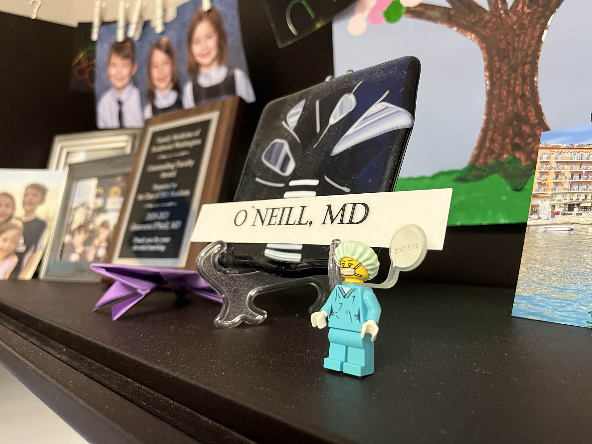 A Lego minifigure of a female medical doctor sits on a desk, with family photos of children in the background.