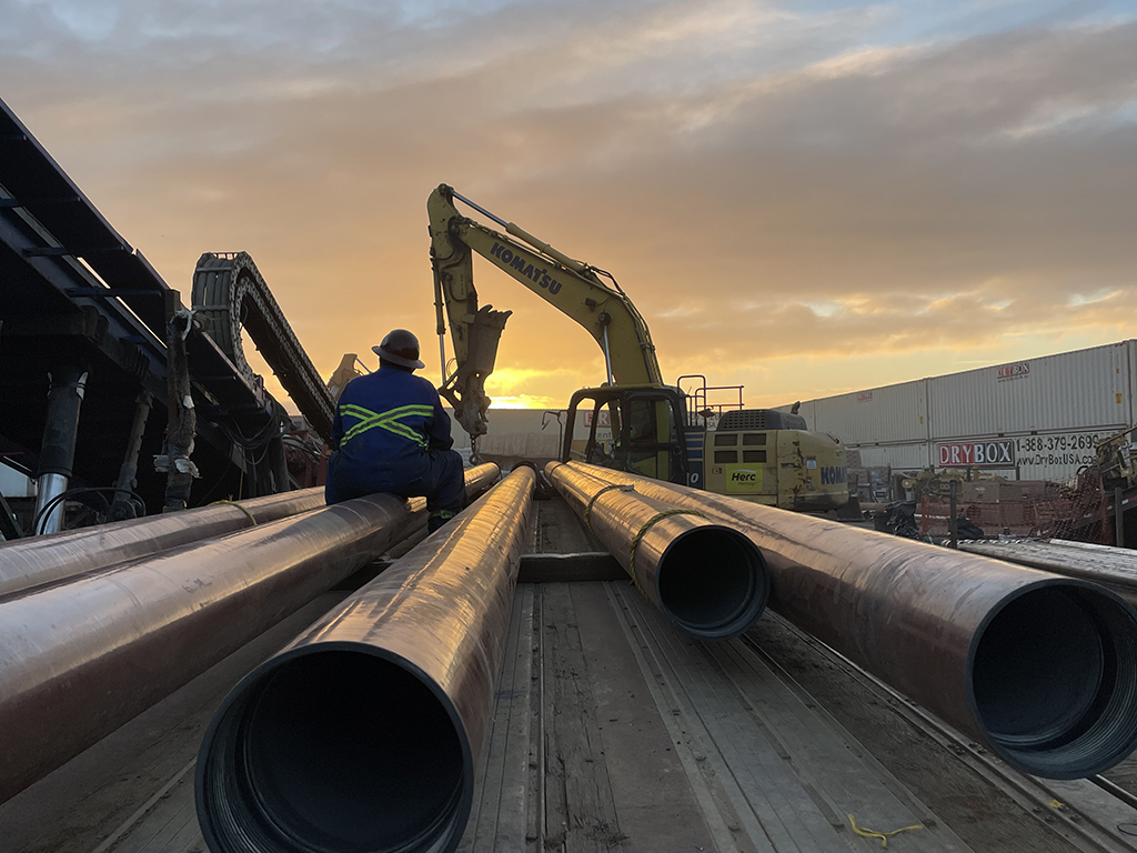A man with workwear and helmet sitting on a stack of long pipes facing the sunset on a construction site.